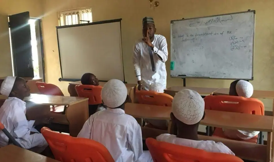 A teacher at a whiteboard of Arabic script with pupils in white kaftans and caps at their desks