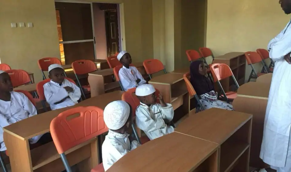 A classroom of pupils at wooden desks in white kaftans and caps, with a teacher standing to the right