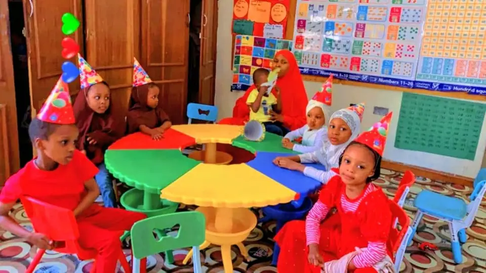 Nursery pupils in red outfits and party hats seated around a colourful round table with their teacher, beside number charts on the wall