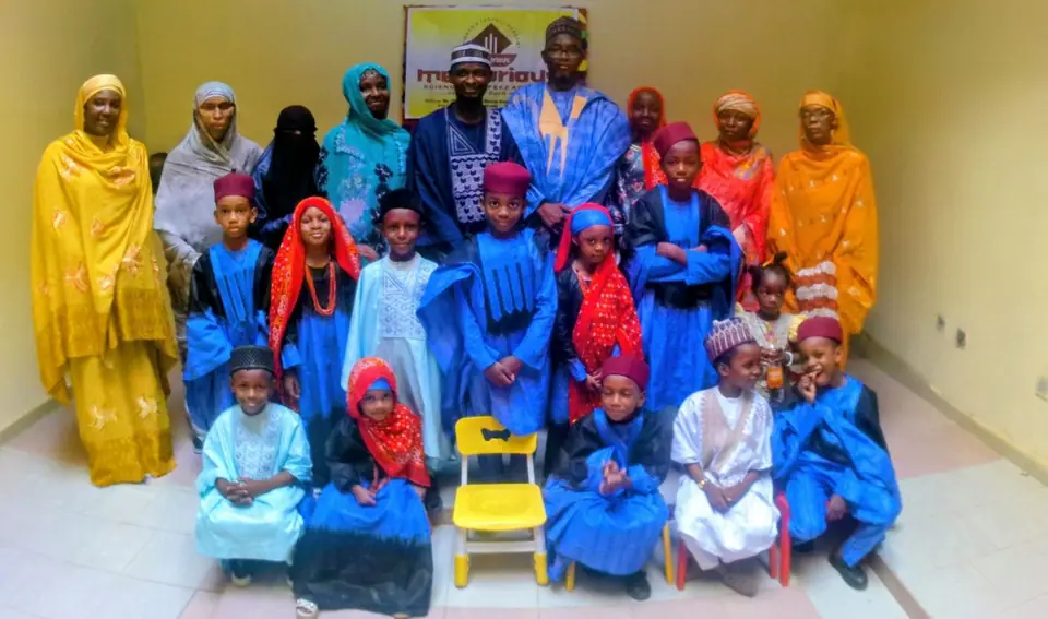 Pupils in blue Hausa dress seated and standing with teachers and staff in front of the Meritorious banner