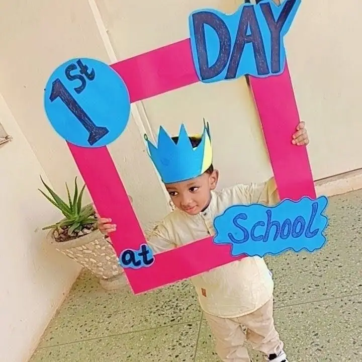 A nursery pupil in a paper crown holding up a pink frame that reads 1st day at school