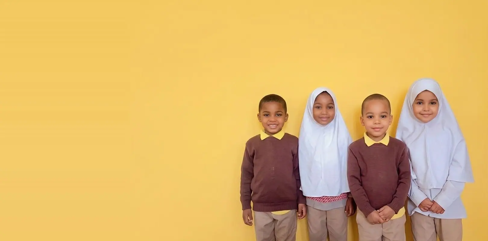Four pupils in school uniform, two boys in maroon jumpers and two girls in white hijabs, standing against a yellow wall