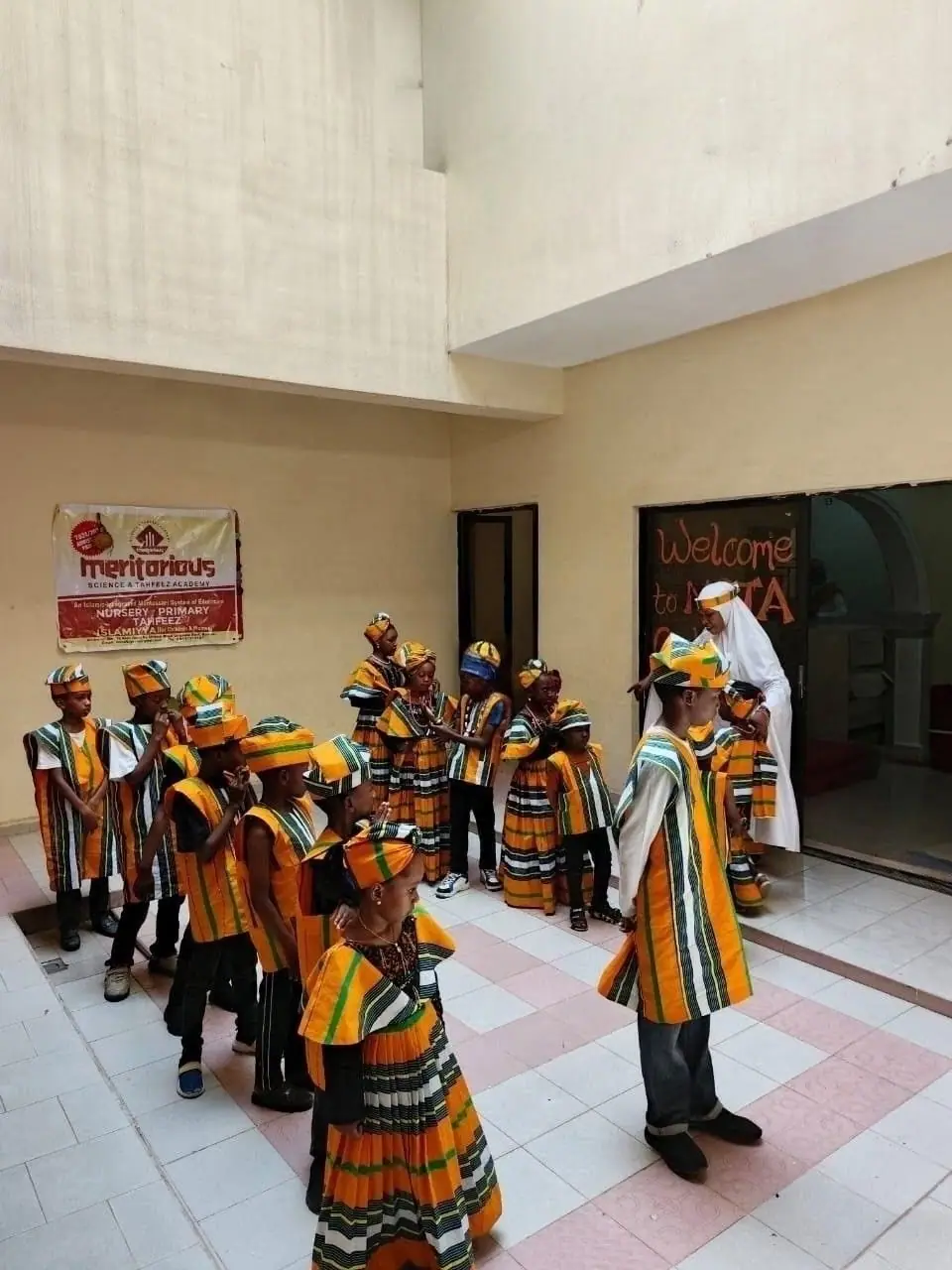 Pupils in cultural day outfits gathering in the school courtyard beside the Meritorious banner and a door painted Welcome to MSTA